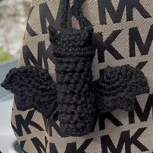 May include: A black crochet bat charm with outstretched wings, hanging from a black cord. The bat is attached to a tan and black patterned handbag with the letters "MK" repeated across the surface.
