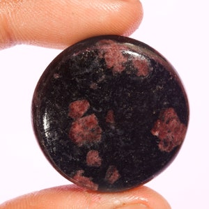 Op de afbeelding: Een ronde, gepolijste edelsteen cabochon met een diepzwarte basiskleur en verspreide, onregelmatige insluitingen van donkerrode en kastanjebruine mineraalafzettingen over het oppervlak, wat zorgt voor een contrastrijk, gespikkeld uiterlijk.