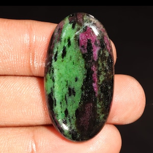 May include: An oval-shaped polished gemstone featuring a vibrant mix of deep magenta-red, bright green, and dark black inclusions. The stone has a smooth, reflective surface with a high-gloss finish, showcasing natural color variations throughout.