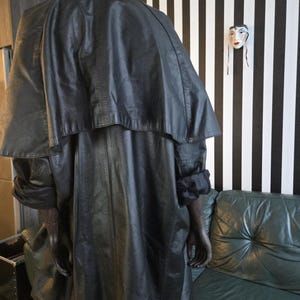 May include: A black leather coat with a cape-like design, displayed on a mannequin. The coat has a long length and a textured appearance. The mannequin is positioned in front of a black and white striped wall.