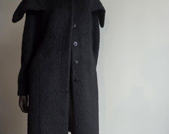 May include: A long, black coat with a unique collar design. The coat features a textured fabric, a button-up front, and a knee-length hem. The coat is displayed on a mannequin.