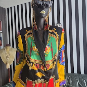 May include: A black mannequin wearing a colorful blazer with gold buttons and a decorative print. The blazer features a mix of yellow, red, green, and gold patterns. The mannequin also wears sunglasses and a gold beaded headpiece.