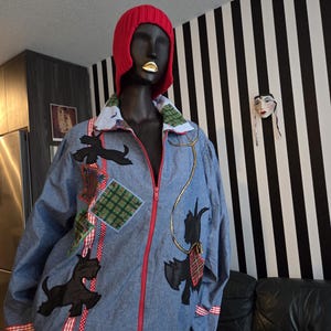 May include: A denim jacket with a red zip and patches of black Scottie dogs and tartan fabric. The jacket has red and white striped trim and is displayed on a mannequin wearing a red knitted hat.
