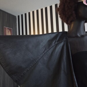 May include: A black leather jacket with a unique wing-like design. The jacket is draped over a mannequin, showcasing its shape and texture. The background features a black and white striped wall.