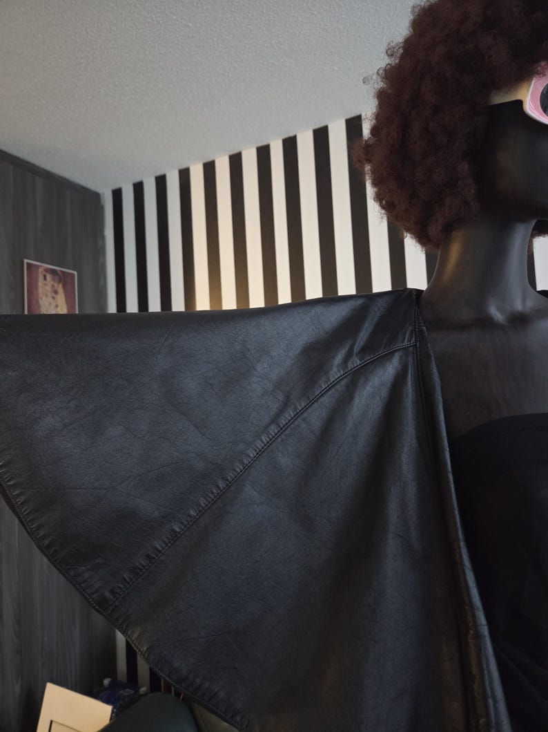 May include: A black leather jacket with a unique wing-like design. The jacket is draped over a mannequin, showcasing its shape and texture. The background features a black and white striped wall.