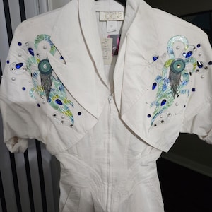 May include: White jumpsuit with a zip-up front and short sleeves. The jumpsuit has a decorative design on the shoulders with blue, green, and silver accents. The design features rhinestones and beads.