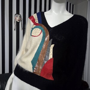 May include: A black and cream-colored sweater with a V-neck design. The left side features a colorful abstract design with red, blue, and brown accents. The sweater has a black ribbed hem and cuffs.