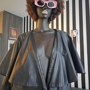 May include: A black leather cape-style coat on a mannequin with a brown afro wig and pink sunglasses. The mannequin is set against a black and white striped wall. The coat has a layered design and a button closure.