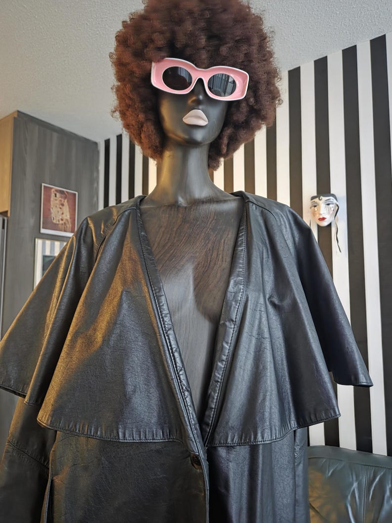 May include: A black leather cape-style coat on a mannequin with a brown afro wig and pink sunglasses. The mannequin is set against a black and white striped wall. The coat has a layered design and a button closure.