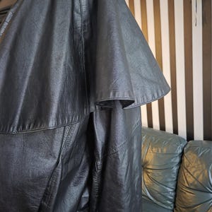 May include: A black leather jacket with a unique cape-like sleeve design. The jacket has a textured appearance and visible stitching details. The garment is displayed against a backdrop of a striped wall and a leather sofa.
