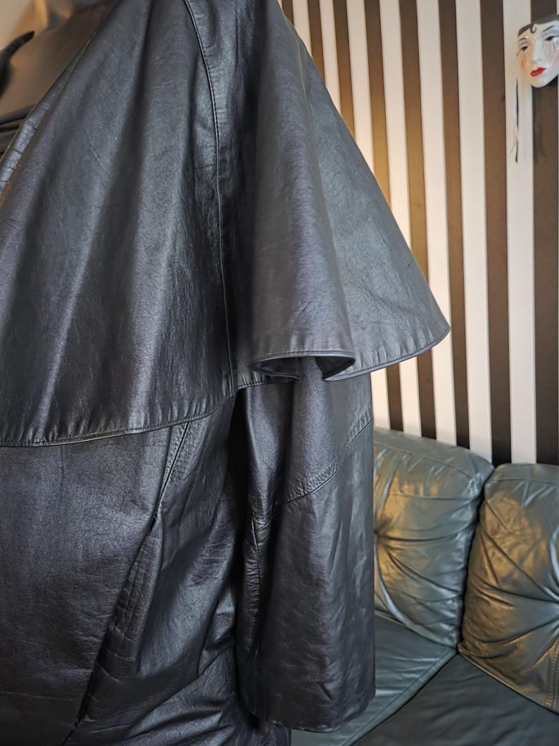 May include: A black leather jacket with a unique cape-like sleeve design. The jacket has a textured appearance and visible stitching details. The garment is displayed against a backdrop of a striped wall and a leather sofa.