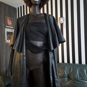 May include: A black mannequin wearing a black leather coat, a sheer black top, and a black bodysuit. The mannequin has a brown afro wig and pink sunglasses. The background features a black and white striped wall and a green leather couch.