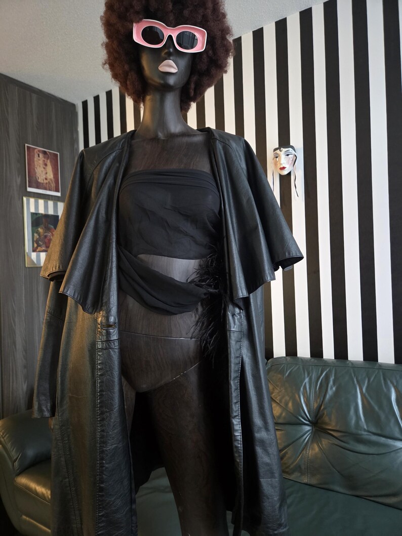 May include: A black mannequin wearing a black leather coat, a sheer black top, and a black bodysuit. The mannequin has a brown afro wig and pink sunglasses. The background features a black and white striped wall and a green leather couch.