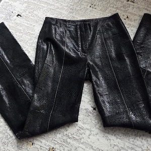 May include: Black faux leather trousers with a snakeskin pattern. The trousers have a high waist and a flared leg. The material has a shiny finish. The trousers are laying on a patterned rug.