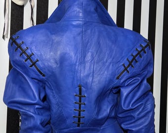 May include: A vibrant blue leather jacket with black stitching details. The jacket features a wide collar and structured shoulders. The back view showcases the intricate stitching along the seams and sleeves. A decorative mask hangs in the background.