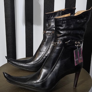 May include: A pair of black leather ankle boots with pointed toes and high heels. The boots have decorative metal accents and a tag that reads "BLAQ BELLA". The boots are displayed on a small, round table.