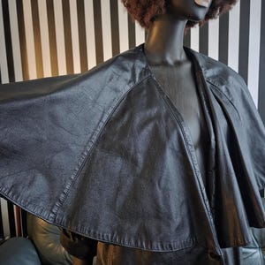May include: A black faux leather cape-style jacket with a wide collar and button closure. The jacket is displayed on a mannequin with a dark complexion, wearing pink sunglasses and a brown afro wig. The background features black and white vertical stripes.