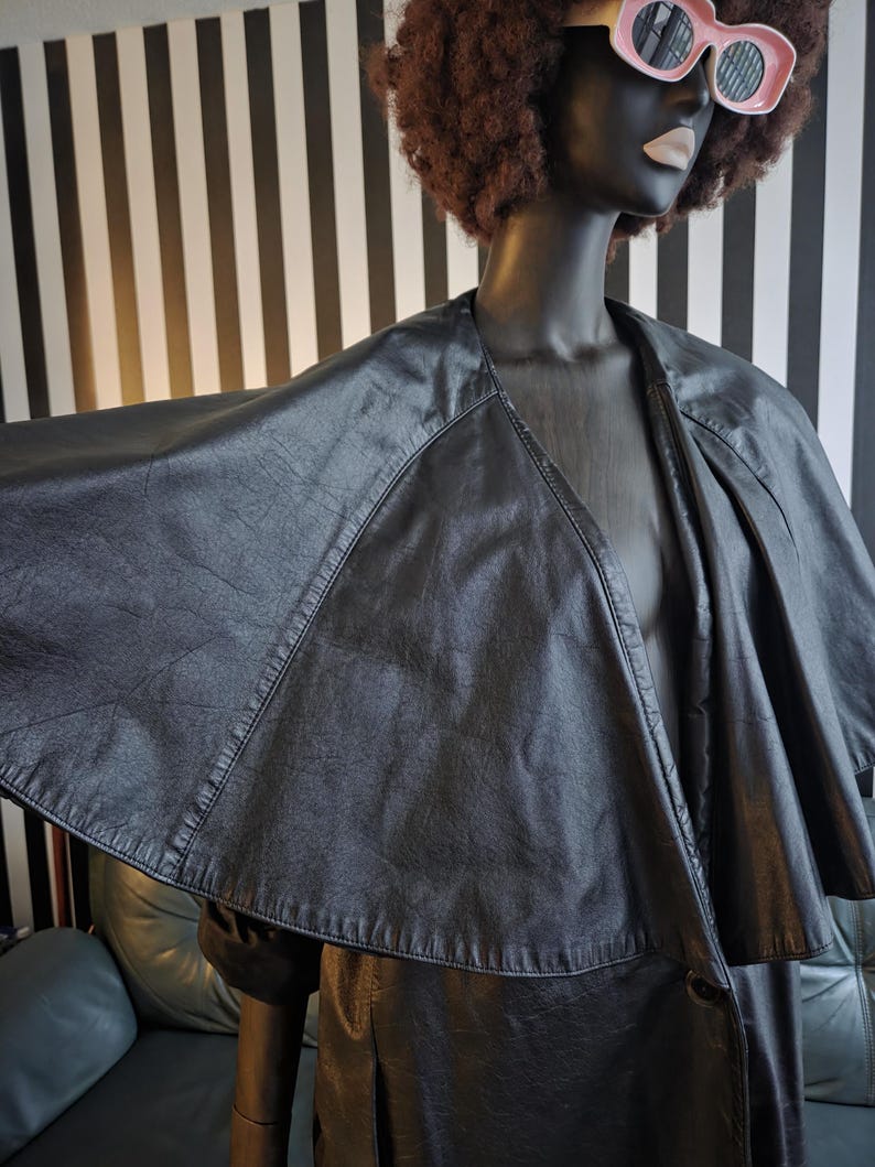 May include: A black faux leather cape-style jacket with a wide collar and button closure. The jacket is displayed on a mannequin with a dark complexion, wearing pink sunglasses and a brown afro wig. The background features black and white vertical stripes.