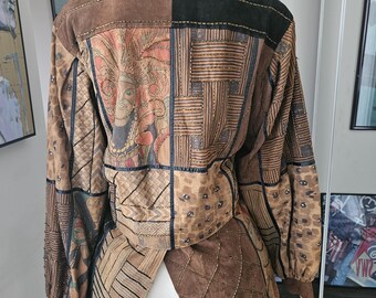 May include: A brown, black, and beige suede jacket with a patchwork design. The jacket has a collar and long sleeves. The back of the jacket features a large, colorful print of a person's face.