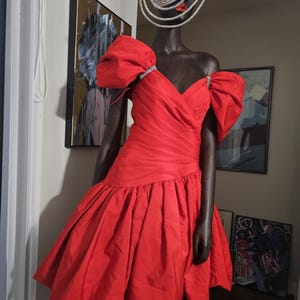 May include: A red, short, puffy-sleeved dress with a sweetheart neckline. The dress is on a mannequin wearing a silver sparkly hat.