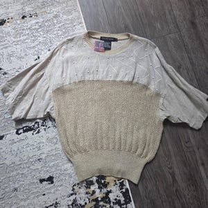 Puede incluir: Jersey beige de manga corta con una sección inferior de punto texturizado y una sección superior lisa. La sección superior presenta bordados blancos en zigzag y pequeños detalles de perlas. La prenda tiene cuello redondo y corte holgado.