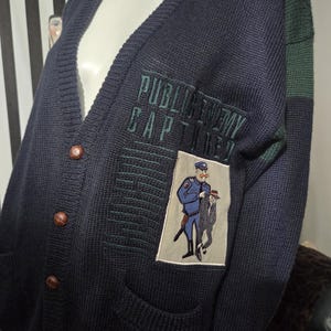 May include: Navy blue cardigan sweater with a V-neck, featuring a patch with a cartoon illustration of a police officer and a man in a suit. The sweater has brown buttons and the text "PUBLIC ENEMY CAPTURED" embroidered on the chest.