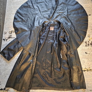 May include: A black leather coat with a unique cape-like collar and a long, flowing design. The coat is open, revealing a dark lining. The garment appears to be vintage, with a classic and dramatic style.