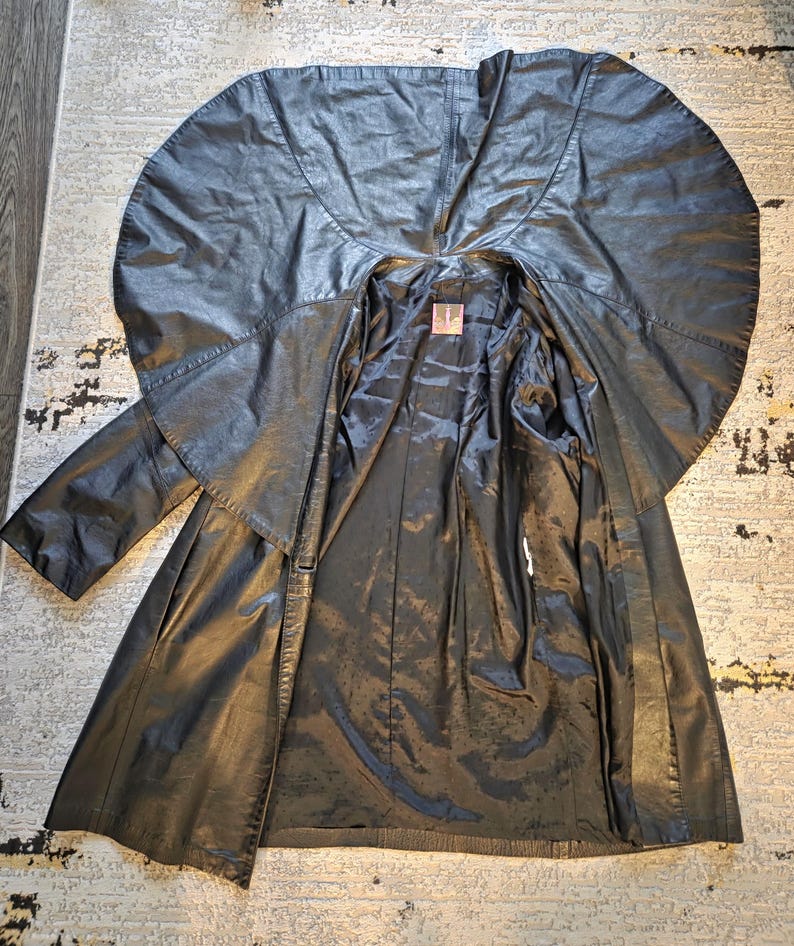 May include: A black leather coat with a unique cape-like collar and a long, flowing design. The coat is open, revealing a dark lining. The garment appears to be vintage, with a classic and dramatic style.