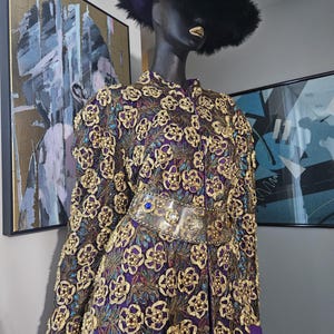May include: A long purple coat with gold and blue floral embroidery. The coat has a clear plastic belt with gold and blue embellishments. The coat is worn by a mannequin with a black fur hat.