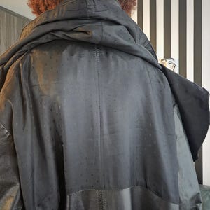 May include: Black hooded coat with a textured, dotted pattern on the upper back and a leather lower section. The coat has a large hood and appears to be worn by a person with curly brown hair. The background features a black and white striped wall.