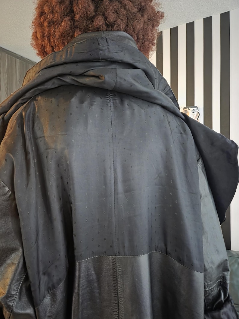 May include: Black hooded coat with a textured, dotted pattern on the upper back and a leather lower section. The coat has a large hood and appears to be worn by a person with curly brown hair. The background features a black and white striped wall.