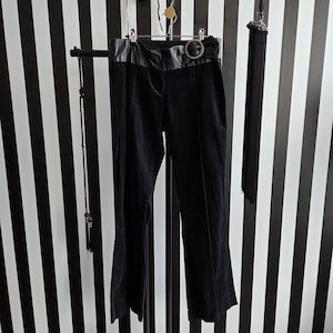 May include: Black velvet trousers with a wide leather waistband and a decorative buckle. The trousers are displayed on a black metal stand against a black and white striped background. A long black tassel hangs to the side.