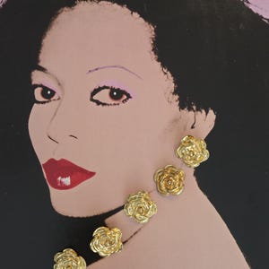 May include: Gold-coloured rose-shaped stud earrings. The earrings are displayed on a background featuring a portrait of a woman with dark hair, red lipstick, and a pink background. The earrings are clustered together.