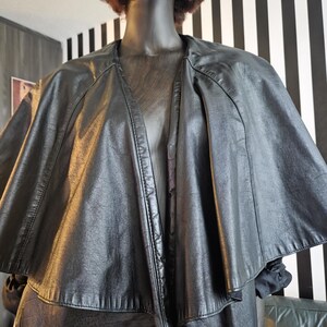 May include: A black leather cape-style jacket with a wide collar and a flowing silhouette. The jacket is displayed on a mannequin with a black face and brown curly hair. The background features black and white striped wall.