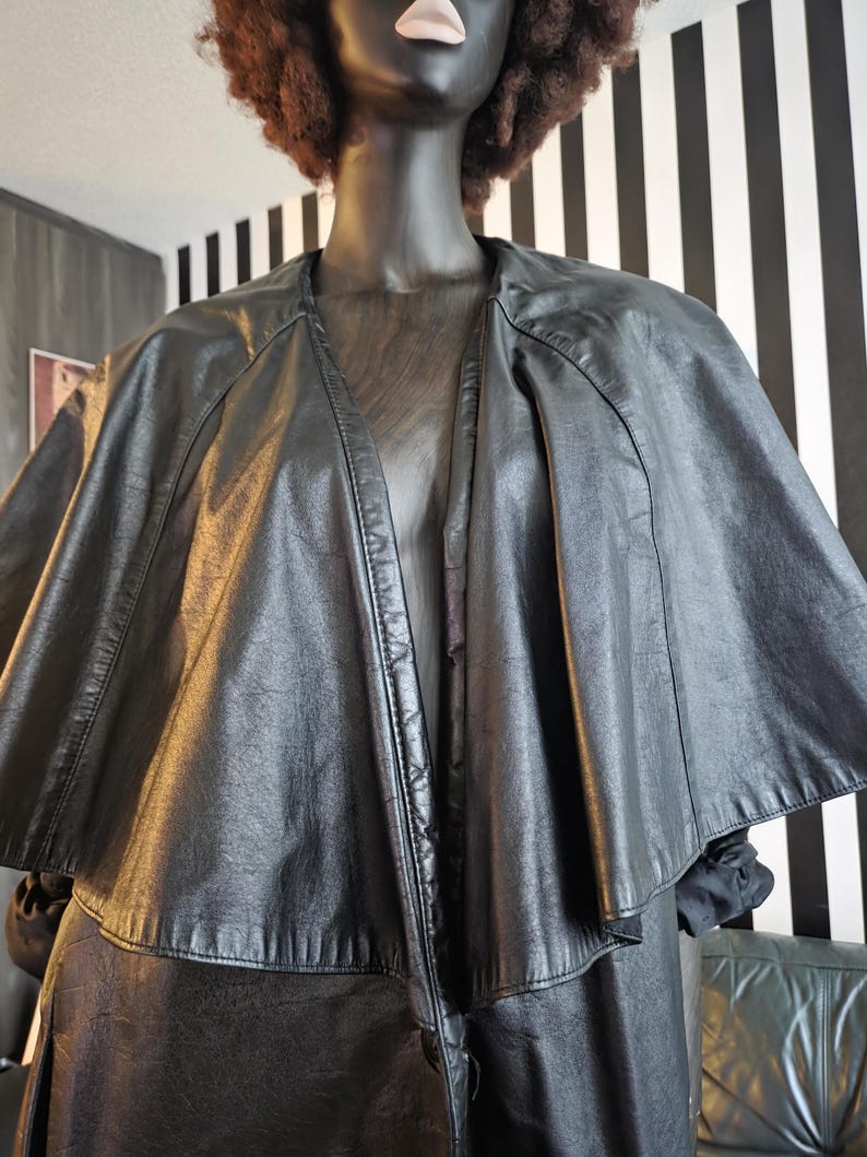 May include: A black leather cape-style jacket with a wide collar and a flowing silhouette. The jacket is displayed on a mannequin with a black face and brown curly hair. The background features black and white striped wall.