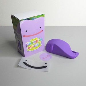May include: A purple Ditto-themed mouse box with a matching mouse, sticker, and a mouse-shaped doorstop. The box features the text "Pokopia Mouse" and a smiling face design. The doorstop is a solid purple color.