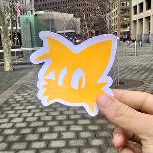 May include: A die-cut sticker featuring a cartoon fox head with two tails, in shades of yellow and white. The sticker has a white border and is held up against a city background.