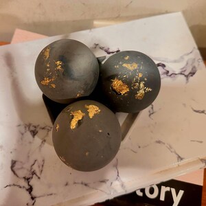May include: Three dark gray spheres with gold leaf accents are arranged in a triangular shape. The spheres are resting on a white and gray marble-patterned surface. The overall aesthetic is modern and minimalist.
