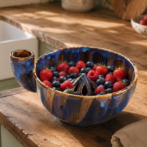 May include: A ceramic citrus juicer bowl with a deep blue and tan glazed finish, featuring a central reamer and a side handle. The bowl is filled with a mixture of fresh red strawberries and small blue blueberries, showcasing a ribbed exterior texture.