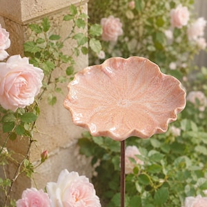 K&ouml;nnte beinhalten: Eine Vogeltr&auml;nke aus Keramik in Form eines gro&szlig;en, gewellten Bl&uuml;tenblatts mit einer gesprenkelten, hellrosa Glasur. Die Schale ist auf einem d&uuml;nnen, dunkelbraunen Metallstab montiert und bietet eine erh&ouml;hte Plattform mit organischer Oberfl&auml;che.