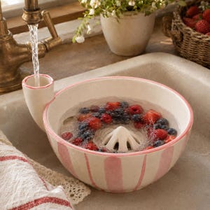 May include: A ceramic berry colander bowl with pink vertical stripes and a pink rim. The bowl features a built-in funnel for water intake and a central reamer-style drain. It is filled with water, fresh red strawberries, and dark blue blueberries.