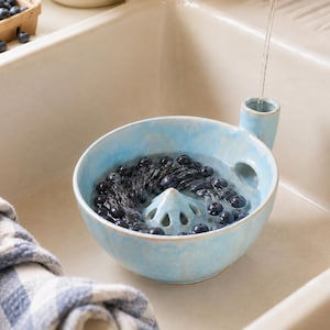 May include: A light blue ceramic berry colander bowl with a central raised cone featuring drainage holes. The bowl has a side handle with a vertical opening and is filled with fresh dark blue blueberries being rinsed under a stream of water.