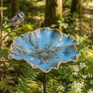 K&ouml;nnte beinhalten: Eine Vogeltr&auml;nke aus Keramik mit einer breiten, gewellten Schale in leuchtendem Blau und dunkelbraunen Sprenkeln. Die Schale ist auf einem d&uuml;nnen Metallstab montiert und wie ein offenes Bl&uuml;tenblatt geformt, das als Becken f&uuml;r V&ouml;gel dient.