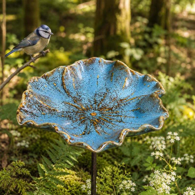 K&ouml;nnte beinhalten: Eine Vogeltr&auml;nke aus Keramik mit einer breiten, gewellten Schale in leuchtendem Blau und dunkelbraunen Sprenkeln. Die Schale ist auf einem d&uuml;nnen Metallstab montiert und wie ein offenes Bl&uuml;tenblatt geformt, das als Becken f&uuml;r V&ouml;gel dient.