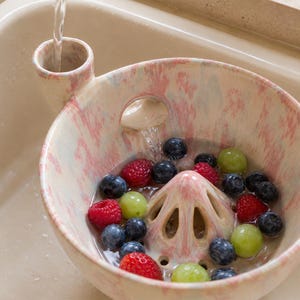 May include: A ceramic berry colander with a pink and white speckled glaze, featuring a central cone-shaped drain and a side funnel for water intake. The bowl is filled with fresh blueberries, raspberries, and green grapes being rinsed with water.