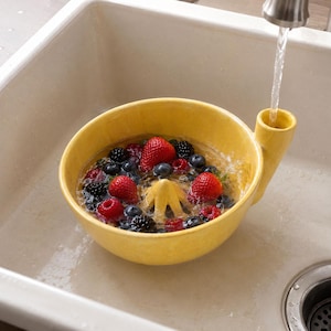 May include: A bright yellow ceramic citrus juicer bowl filled with water and a mix of fresh red strawberries, dark blackberries, and small blueberries. The bowl features a central reamer and a side spout, positioned under a running kitchen faucet.