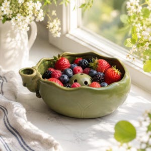 May include: A sage green ceramic bowl shaped like a cabbage leaf, featuring a small integrated handle and a central drainage hole, filled with a mix of fresh red strawberries, raspberries, and dark blue blackberries.