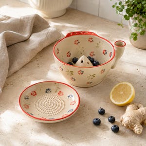 Könnte beinhalten: Ein Zitruspressen-Set aus Keramik, bestehend aus einer cremefarbenen Schale mit zentralem Presskegel und einer passenden flachen Reibeplatte. Beide Teile sind mit kleinen roten und blauen Blumenmustern verziert und haben einen leuchtend roten Rand.