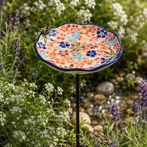 May include: A round, scalloped-edge ceramic bird bath featuring a colourful floral pattern in shades of red, orange, and blue on a cream background, supported by a thin black metal stake, with a small bee resting on the rim of the basin.