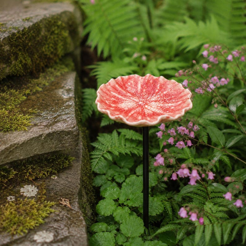 K&ouml;nnte beinhalten: Ein Gartenstecker mit einer runden, gewellten, rot-wei&szlig; gesprenkelten Keramikschale auf einem d&uuml;nnen, geraden schwarzen Metallstab. Die Schale hat eine texturierte, organische Oberfl&auml;che mit einem welligen Rand, &auml;hnlich einem Pilzhut.